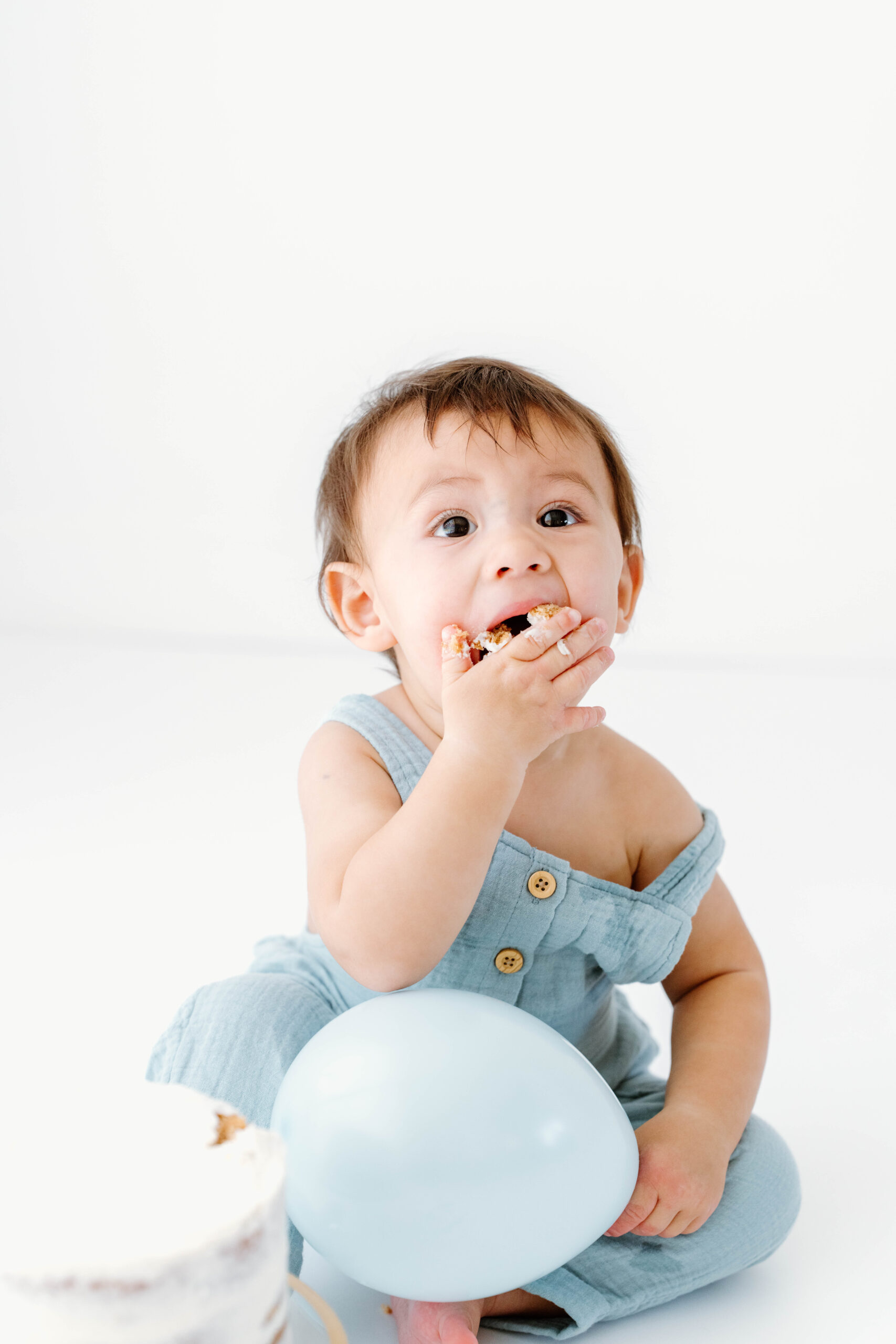 baby eating cake while holding a blue balloon in a natural light studio in southern california.
