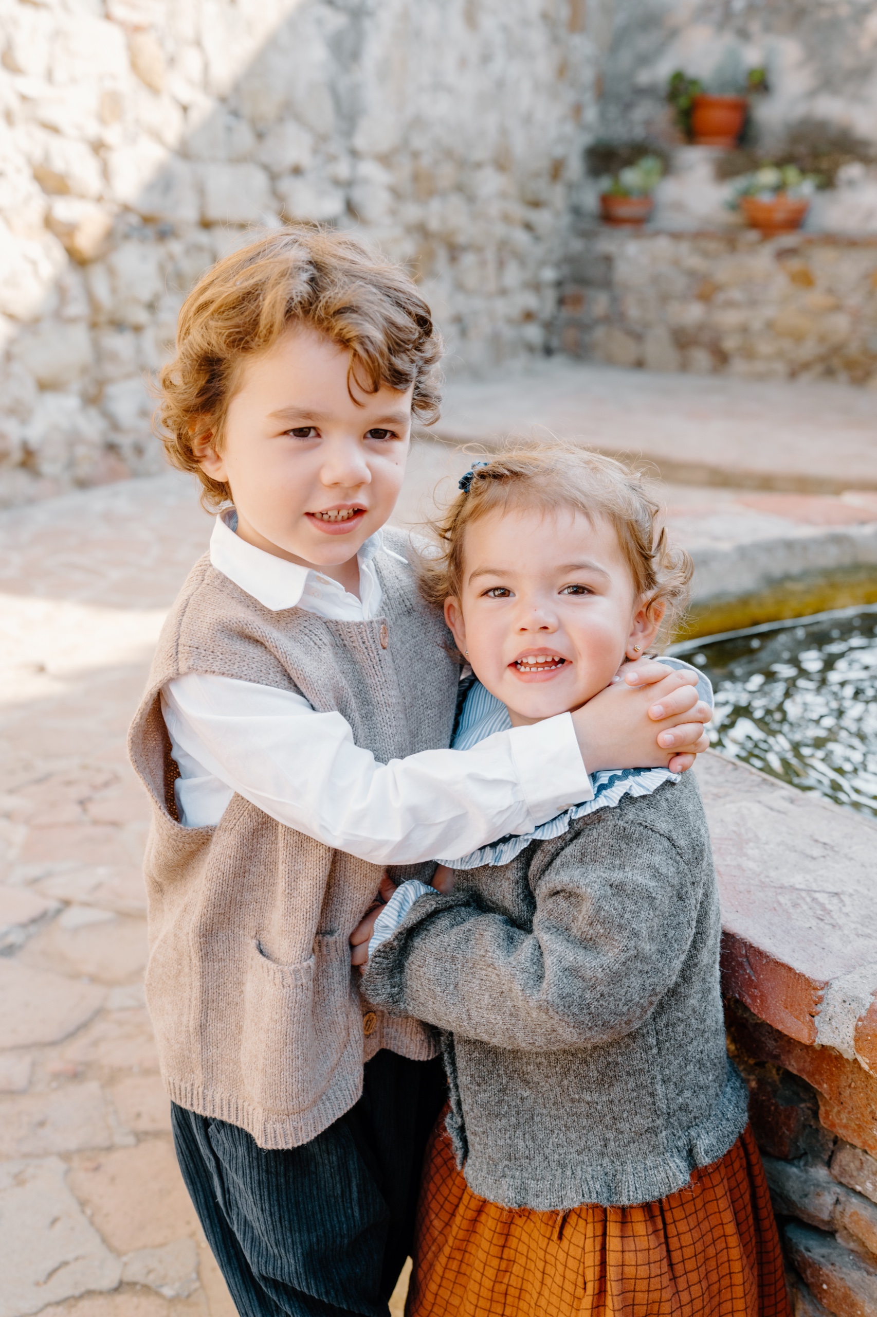 Two young children hugging and smiling beside a fountain at Mission San Juan Capistrano