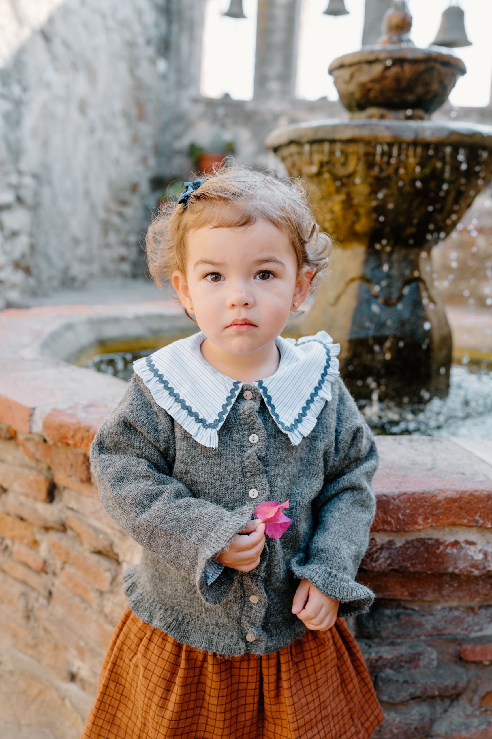 Toddler girl in a grey cardigan holding a pink flower beside a stone fountain