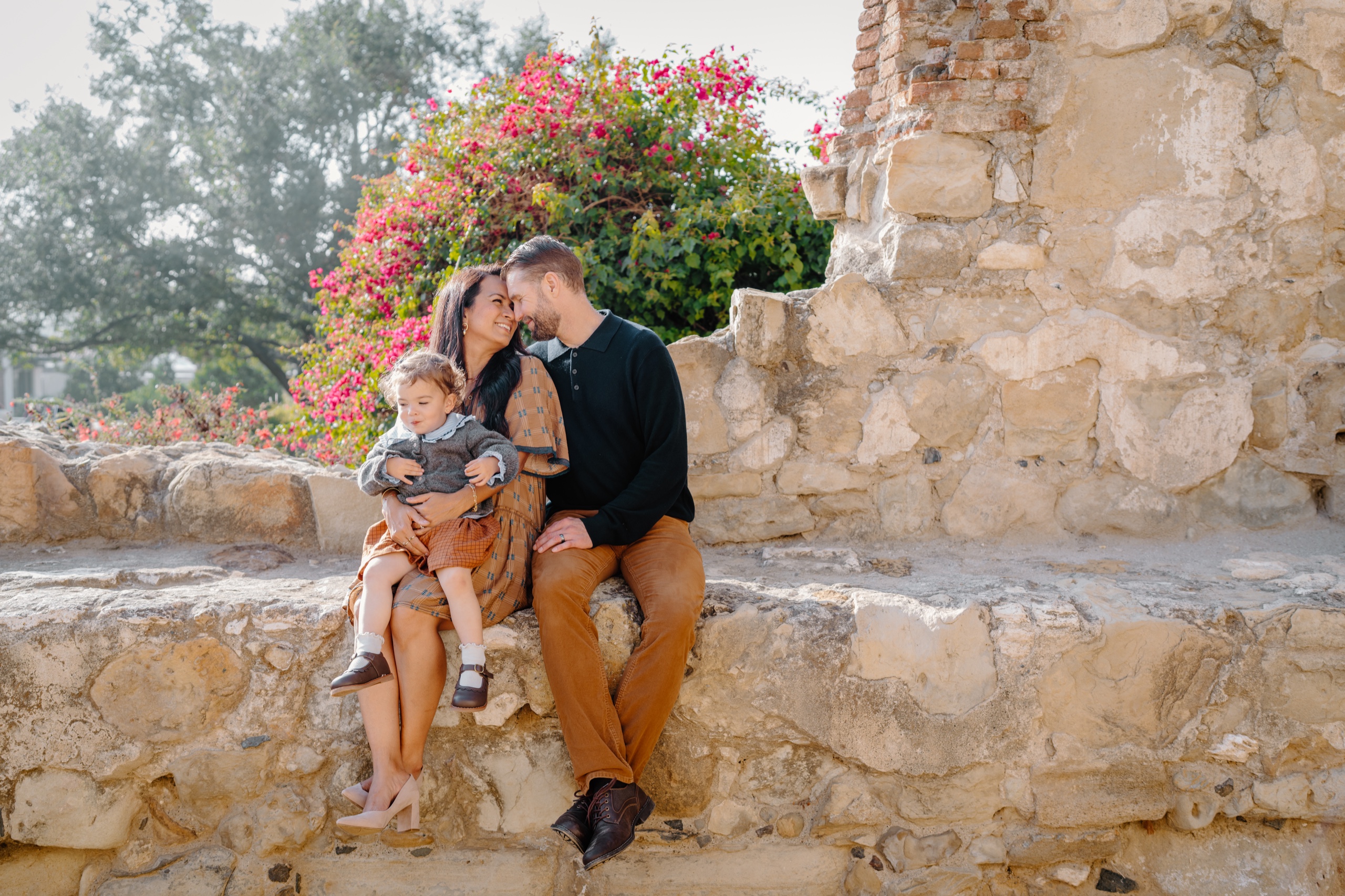 Couple with their toddler sitting on a stone wall under pink bougainvillea