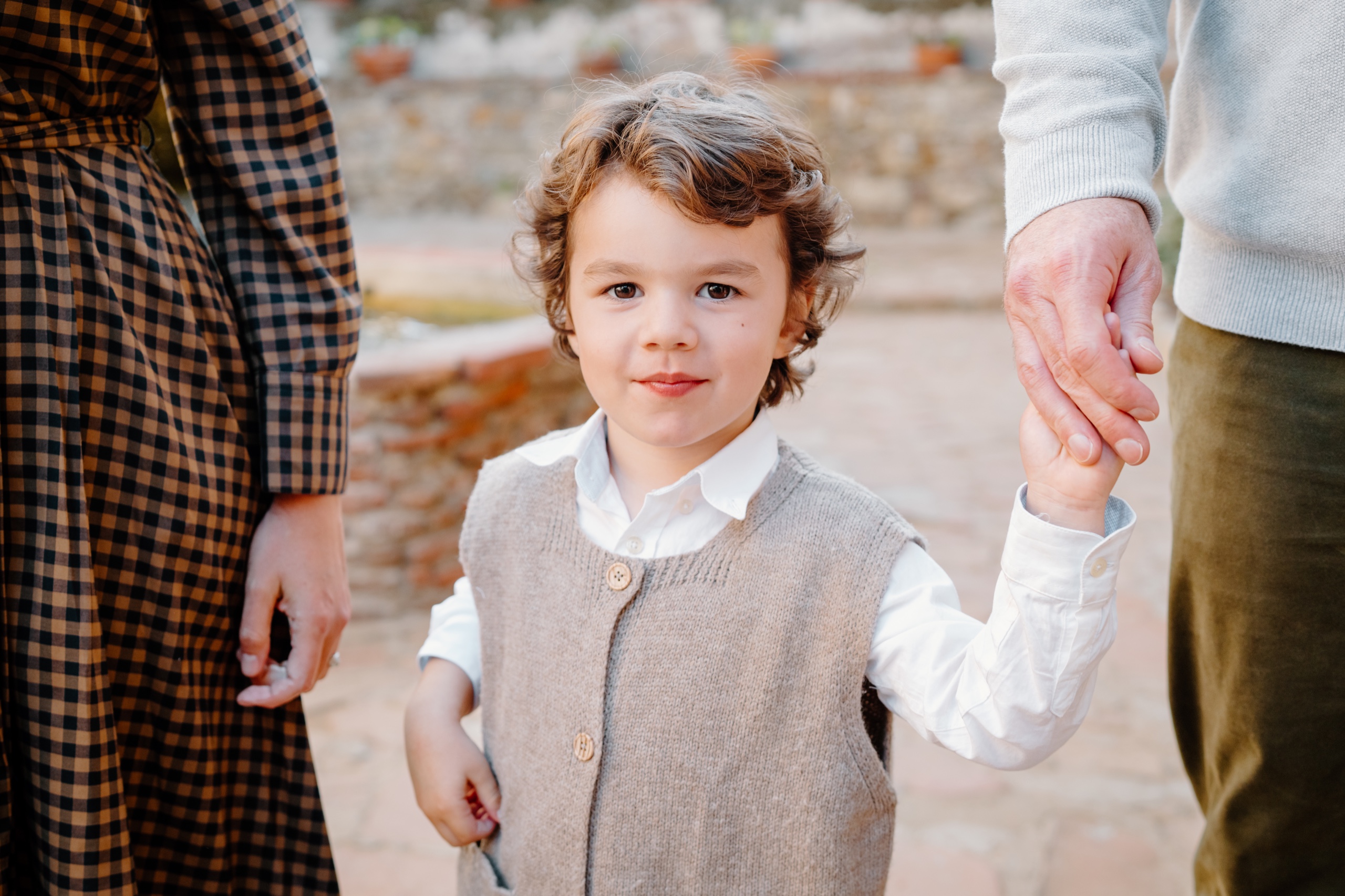 Curly-haired little boy in a knit vest holding a grown-up’s hand