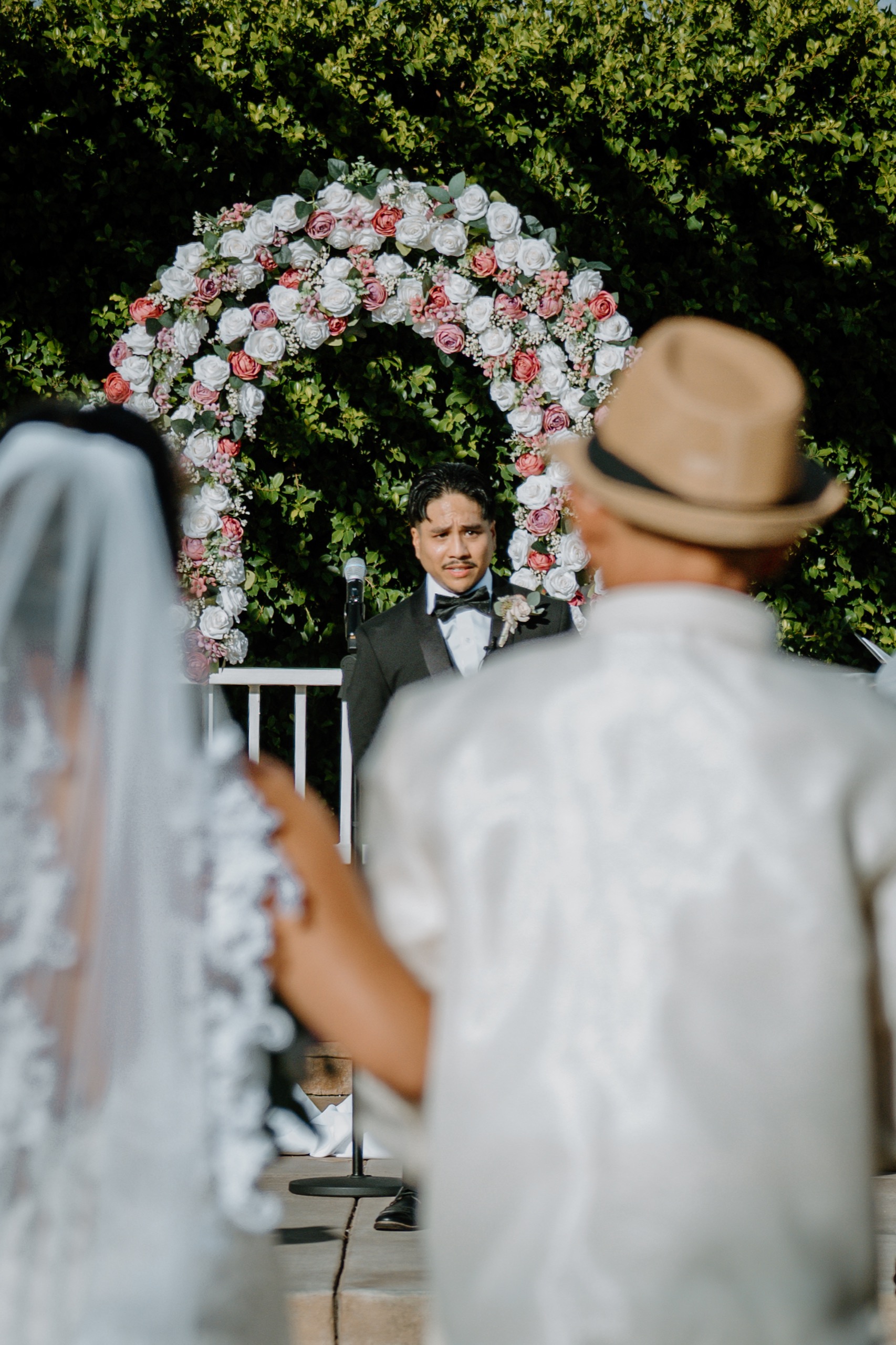 Bride in a lace veil walking toward the ceremony, framed by a white and pink rose arch