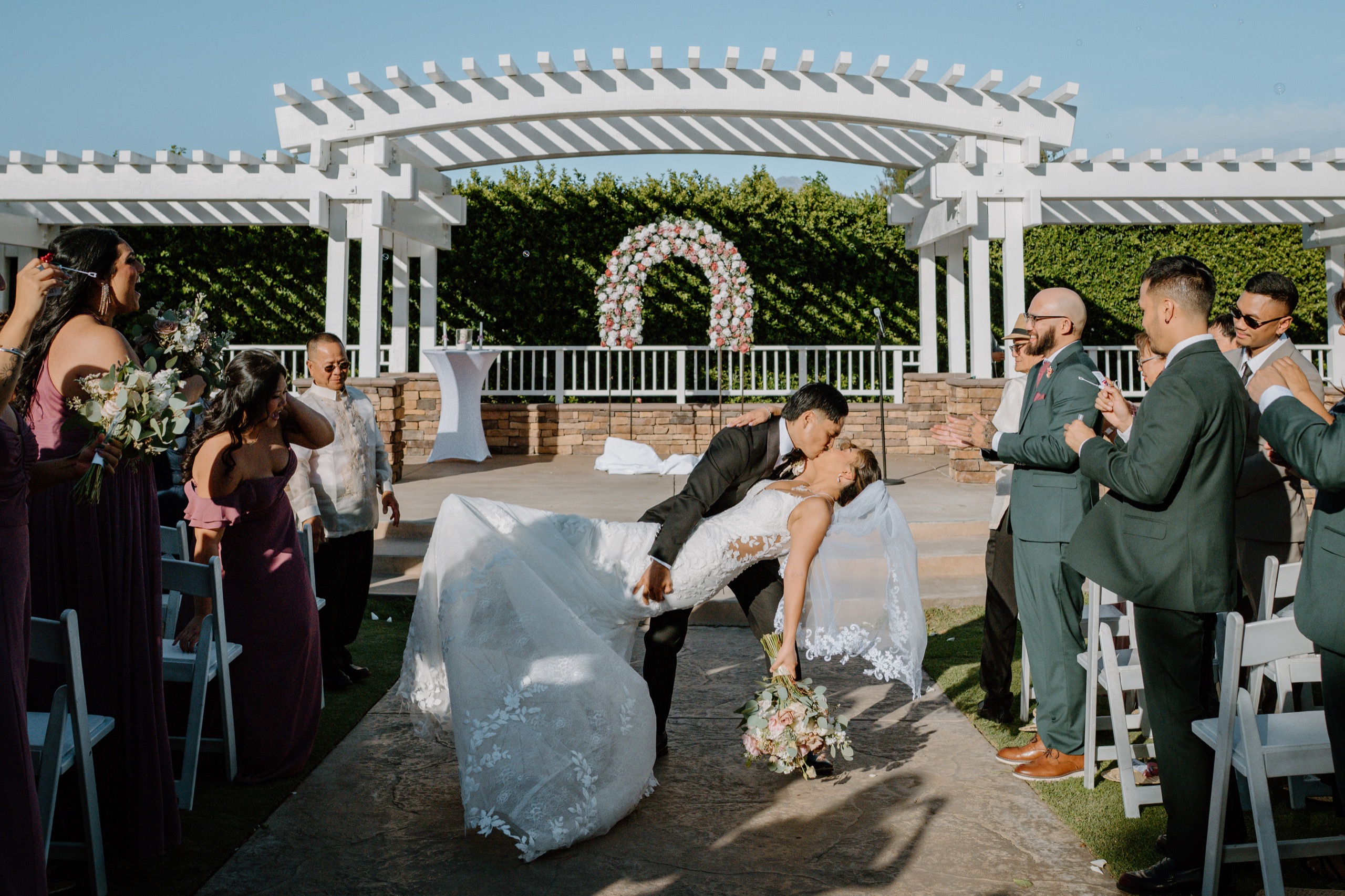 Groom dipping his bride for a kiss in the aisle as guests cheer under a white pergola