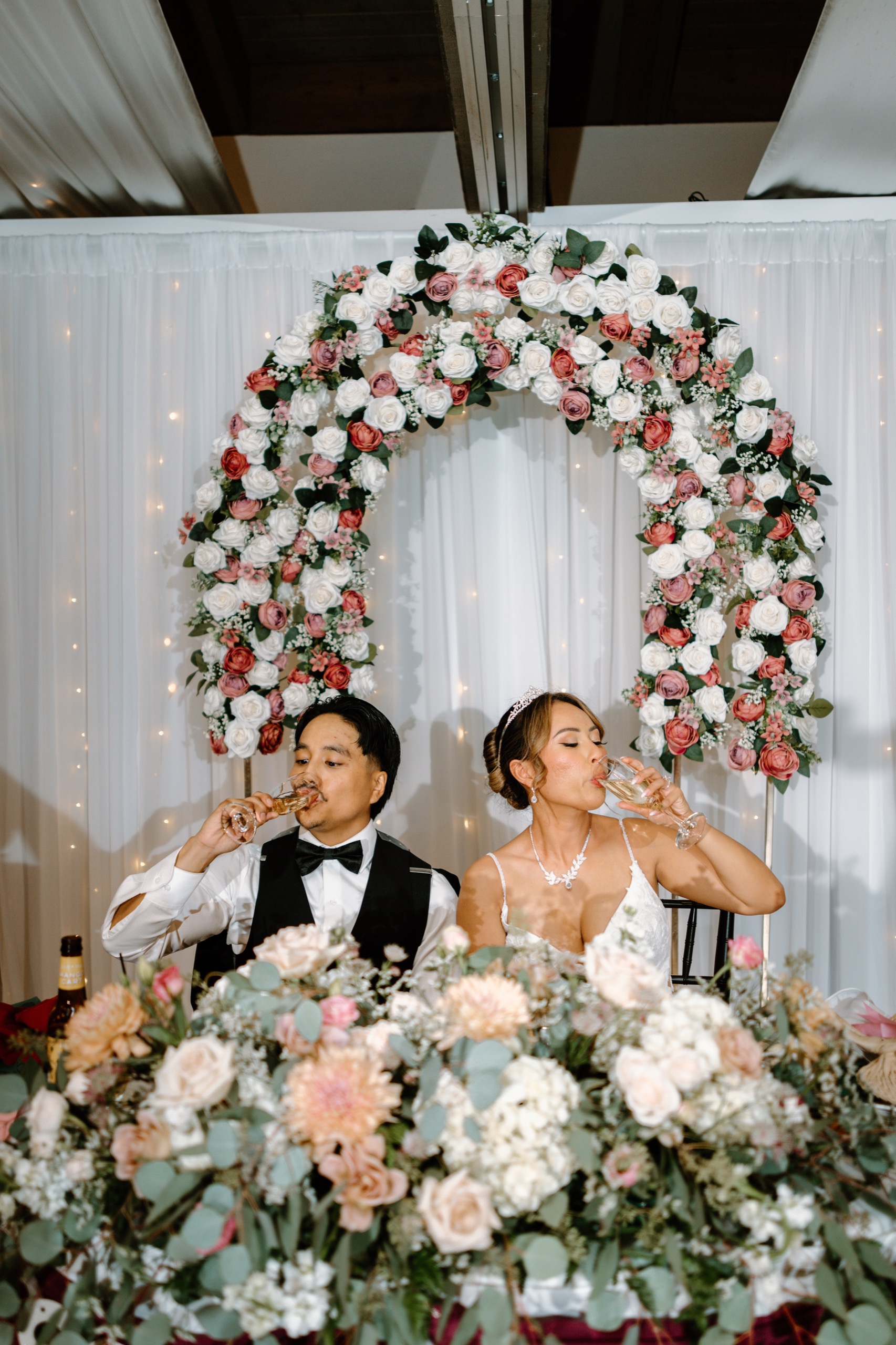 Bride and groom toasting at the sweetheart table in front of a white and pink rose arch